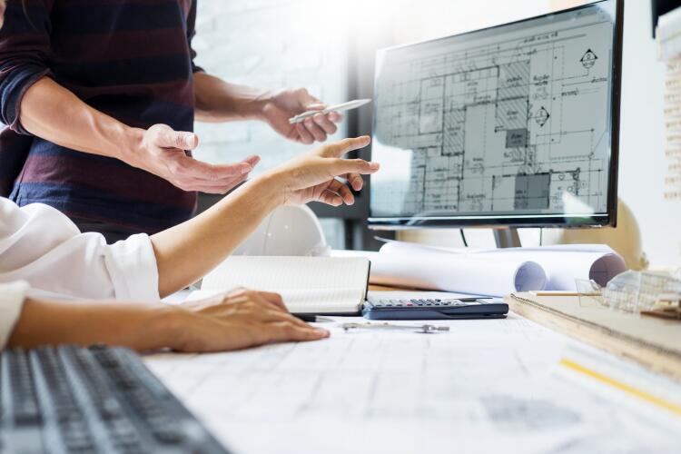 Two engineers are collaborating at a desk filled with blueprints and technical drawings. They are discussing the plans displayed on a computer monitor, which include detailed schematics for the installation of Flowflex plumbing fittings and valves.