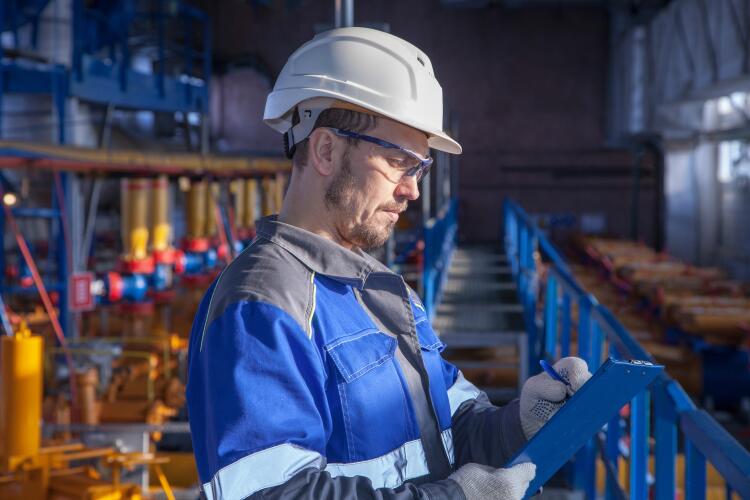 An industrial engineer wearing a white hard hat and safety glasses is working inside a factory. The background features various industrial machinery and equipment, indicating the application of Flowflex products in industrial applications.