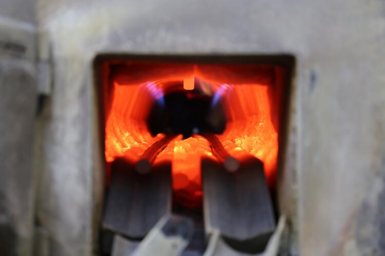 A close-up view inside a high-temperature furnace with glowing orange interior walls and blue flames. Brass billets are being heated inside the furnace, showcasing Flowflex manufacturing.