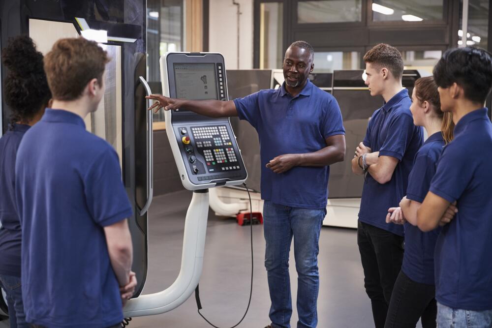 An instructor in a blue polo shirt is explaining a CNC machine to a group of young trainees. This image highlights Flowflex’s commitment to education and training, providing new talent with the skills and knowledge needed for a successful career in precision manufacturing.